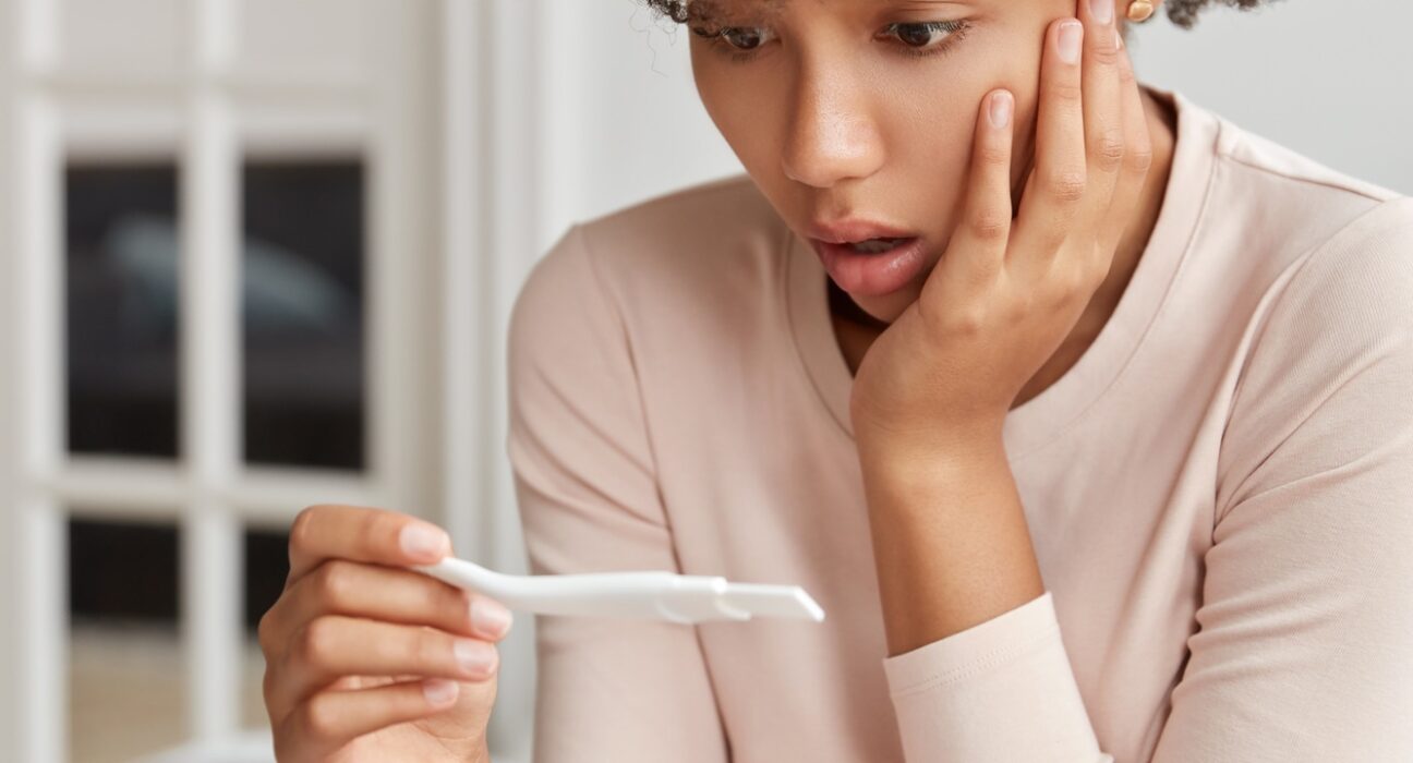 Vertical shot of stupefied woman with Afro haircut, checks positive result on pregnancy test, finds