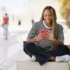 Black girl texting with a smartphone sitting on a bench outdoors, wearing her hair in braids.