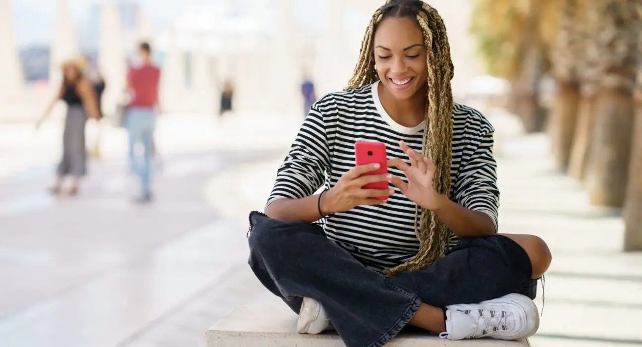 Black girl texting with a smartphone sitting on a bench outdoors, wearing her hair in braids.