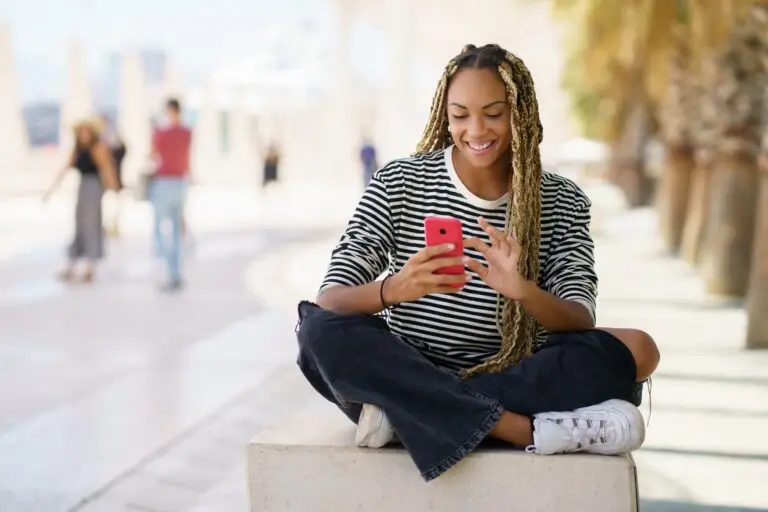 Black girl texting with a smartphone sitting on a bench outdoors, wearing her hair in braids.
