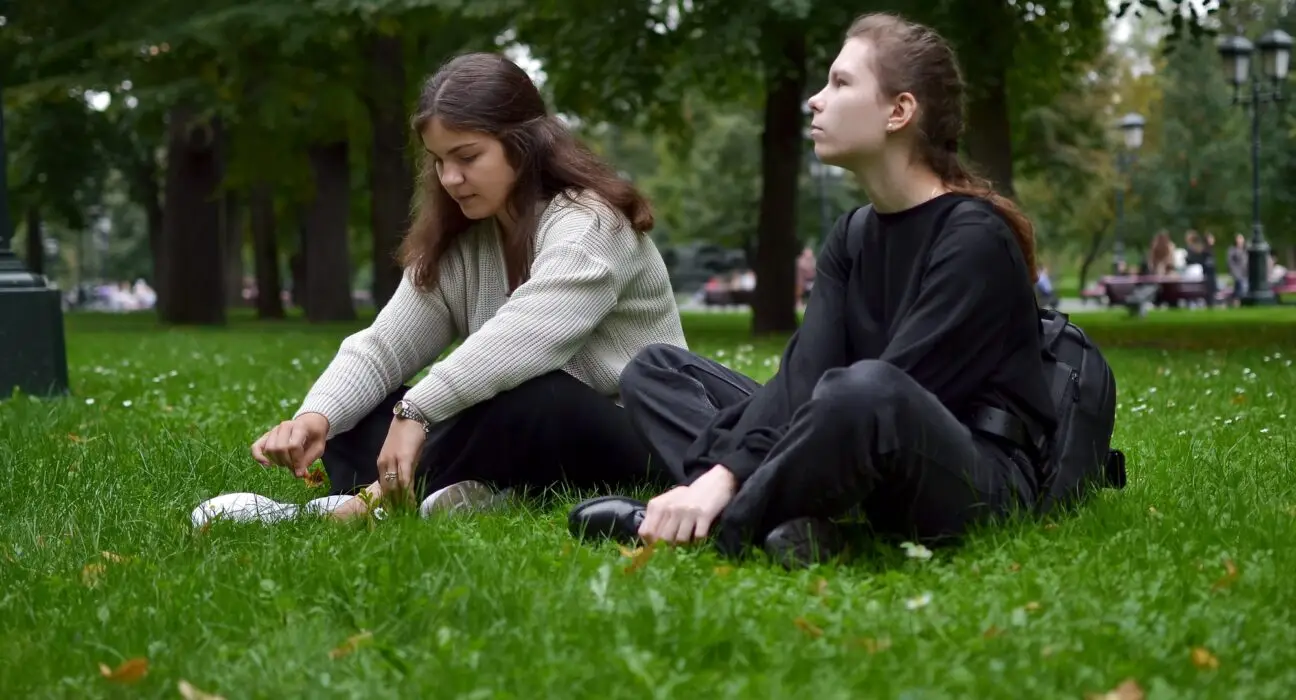 Two young women are sitting on the lawn in the park talking, sharing experiences and victories.