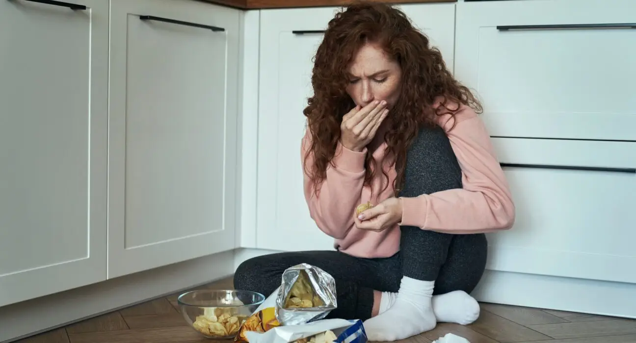 Young caucasian woman eating snacks greedily on the kitchen floor