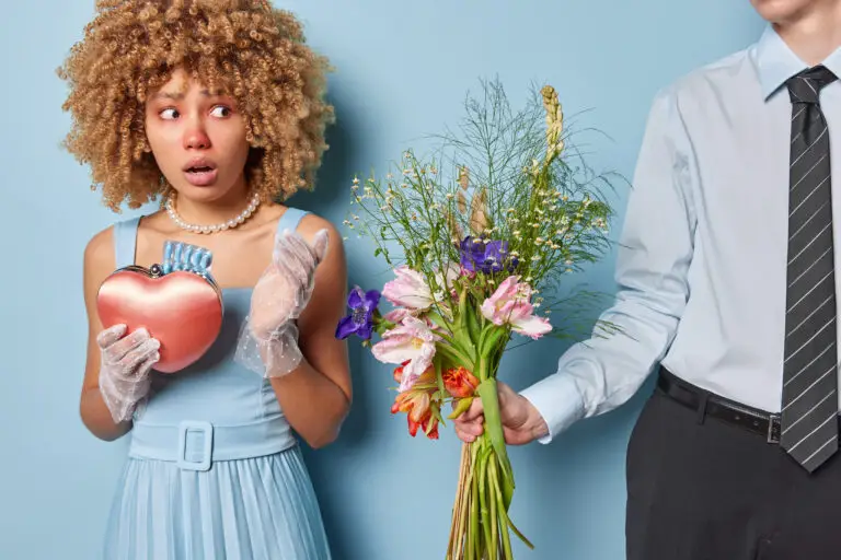 Shocked Curly Haired Woman With Bouquet Of Flowers In Hand Her Boyfriend Expresses His Love Despite Being Aware Of Her Allergies Wears Elegant Clothing Looks Worried Isolated Over Blue Background